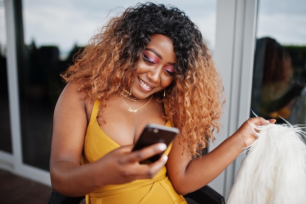 Glamour mujer afroamericana en vestido amarillo y capa de lana blanca sentada en una silla y mirando el teléfono celular