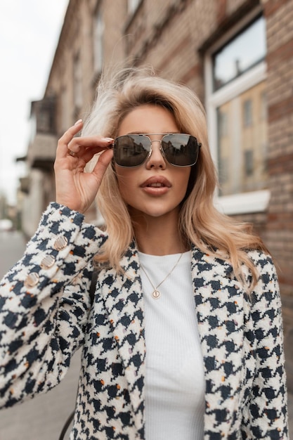 Glamour trendiges Mädchen mit Sonnenbrille in modischer Jacke und weißem T-Shirt geht auf der Straße Eleganter urbaner Look der Frau