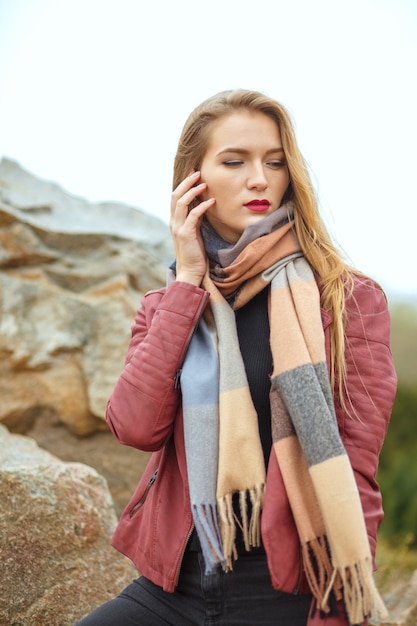 Glamouröse blonde Frau mit Schal und Jacke, die in der Nähe der Felsen posiert