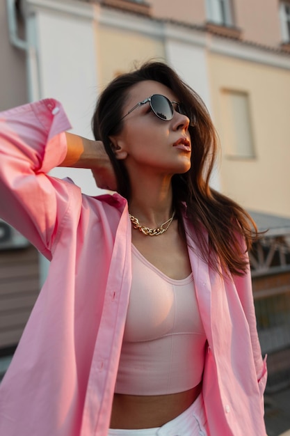 Glamouröse junge sexy Modellfrau mit modischer Vintage-Sonnenbrille und goldener Kette in einem trendigen rosa Hemd und Top, die in der Stadt spazieren gehen