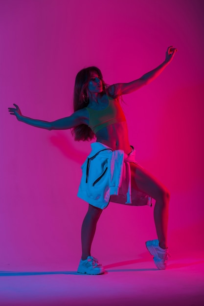 Glamouröse junge Tänzerin in trendiger Sport-Jugendkleidung, die in einem Raum mit mehrfarbigem rosafarbenem Disco-Stil posiert. Attraktives Mädchenmodel steht im Studio mit hellem Neonlicht