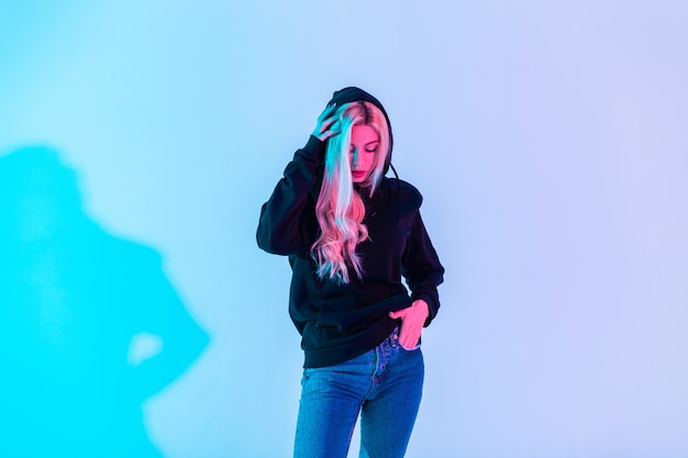 Glamouröse schöne junge blonde Frau in einem modischen schwarzen Sweatshirt trägt eine Kapuze in Blue Jeans auf einem bunten neonrosa Lichthintergrund im Studio