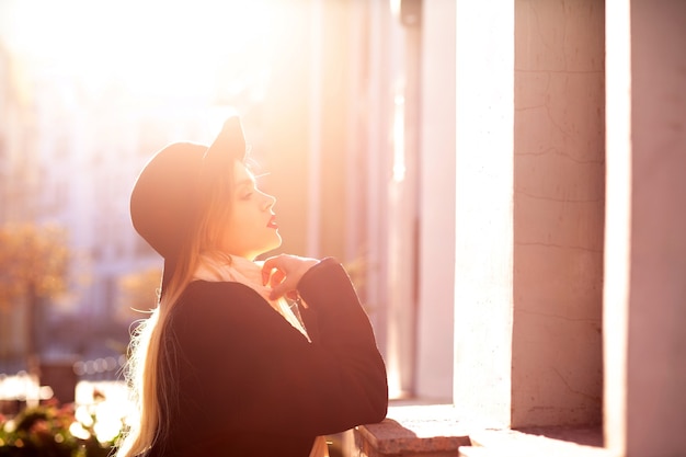 Glamouröses blondes Model mit Mantel und Hut, das an der alten Straße posiert. Platz für Text