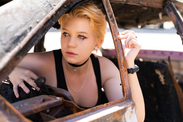 Glamouröses Mädchen in Hosen und kurzen Haaren und Make-up posiert neben einem kaputten alten Auto