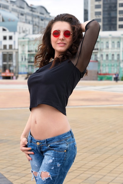 Glamourosa jovem morena de óculos de sol vermelhos redondos, se divertindo na cidade
