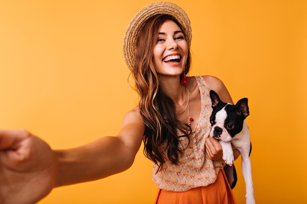 Glamourosa mulher rindo com buldogue francês fazendo selfie em fundo laranja Alegre garota caucasiana se divertindo com seu animal de estimação