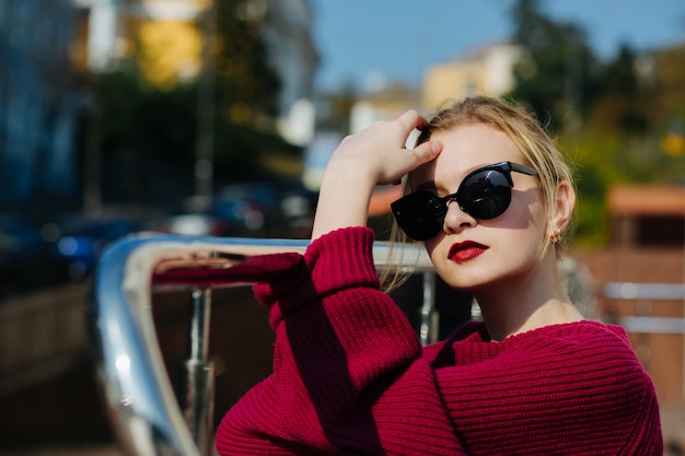 Glamourosa senhora loira de suéter e óculos posando para a cidade. Espaço para texto