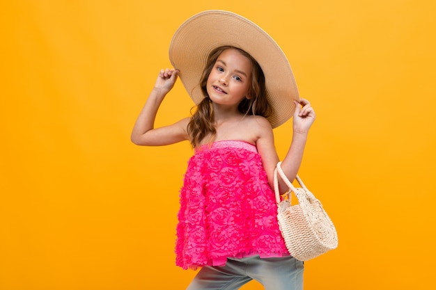 Glamouroso niño encantador con un sombrero de paja y una bolsa redonda en una pared amarilla