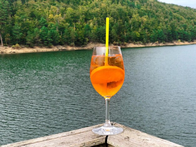 Foto glas aperol-spritz-cocktail mit see und grünem wald im hintergrund