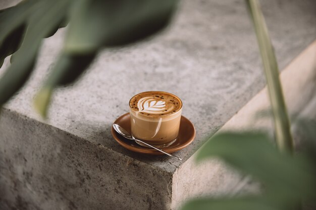 Glas Cappuccino mit Latte Art auf Untertasse und mit Löffel auf Betonhintergrund mit tropischer Pflanze davor