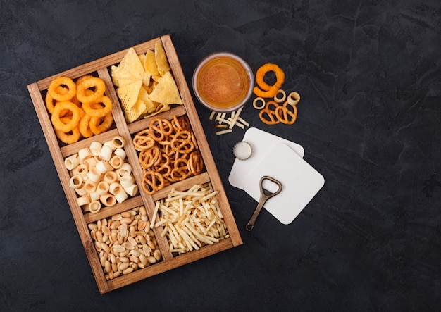 Glas Craft Lagerbier und Öffner mit Snacks auf dunklem Hintergrund. Brezel, salzige Kartoffelsticks, Erdnüsse, Zwiebelringe mit Nachos in Vintage-Box mit Öffnern und Bierdeckeln. Ansicht von oben