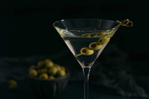 Glas des klassischen trockenen Martini-Cocktails mit Oliven auf dunklem Steintisch vor einem schwarzen Hintergrund.