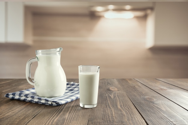 Glas frische Milch und Krug auf hölzerner Tischplatte mit Unschärfeküche als Hintergrund.