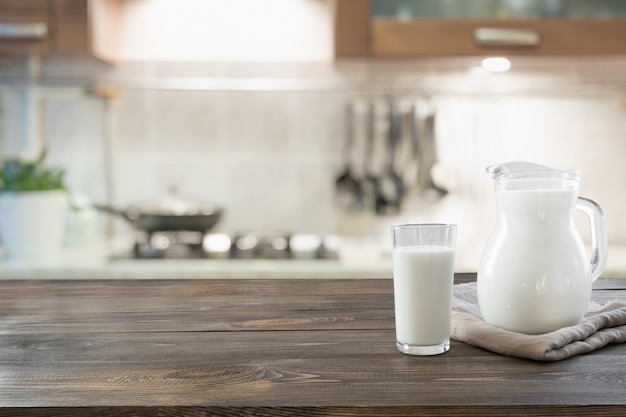 Glas frische Milch und Krug auf hölzerner Tischplatte mit Unschärfeküche als Hintergrund.