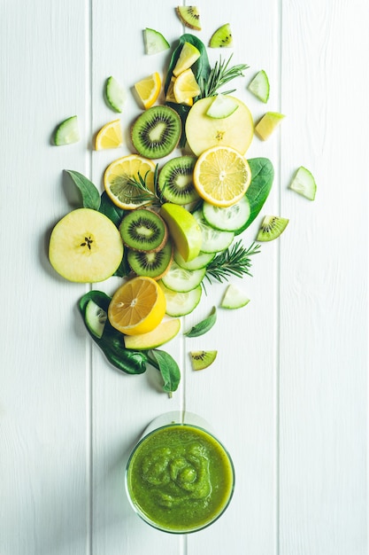 Glas grüner Smoothie mit Gemüse, Obst und Kräutern, aus dem es auf einer weißen Holzoberfläche hergestellt wird