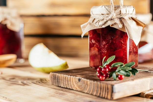 Glas hausgemachte Preiselbeer- und Birnenmarmelade mit Kraftpapier auf dem Deckel auf Holztisch neben frischen Preiselbeeren und Birnen