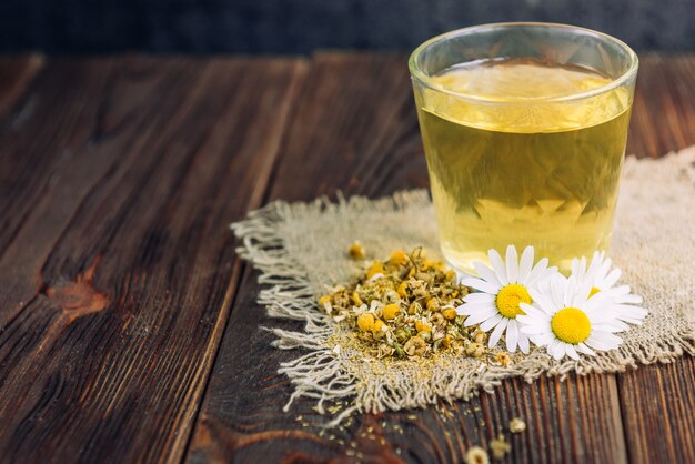 Glas Kräutertee mit Kamillenblüten auf dunklem hölzernem Hintergrund.