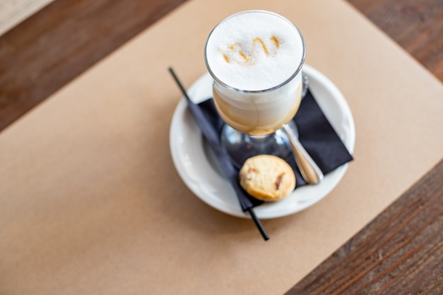 Glas Latte Macchiato mit Löffelstroh und Teller mit Keksen auf einem Holztischköstliche süße Br...