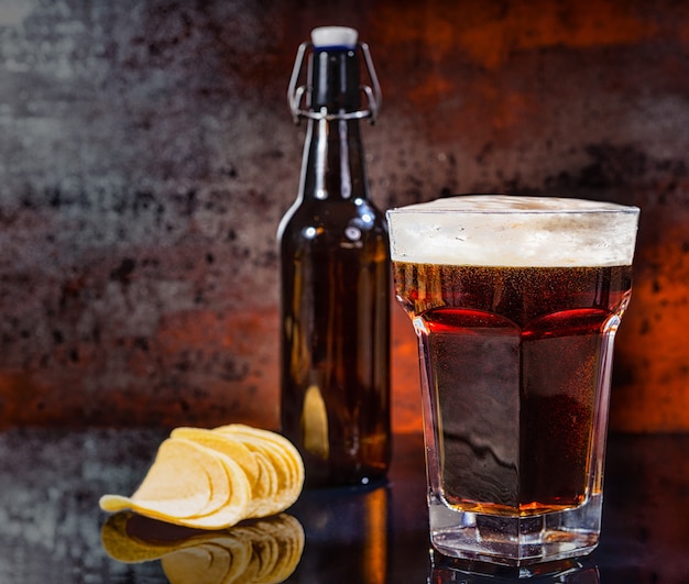 Glas mit frisch gegossenem dunklem Bier, Bierflasche in der Nähe von verstreuten leckeren Pommes auf einer schwarzen Spiegeloberfläche. Lebensmittel- und Getränkekonzept