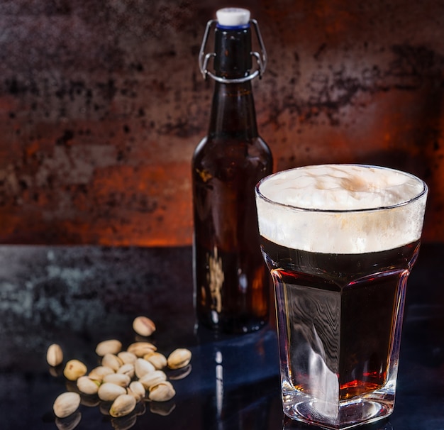 Glas mit frisch gegossenem dunklem Bier, Bierflasche in der Nähe von verstreuten Pistazien auf einer schwarzen Spiegeloberfläche. Lebensmittel- und Getränkekonzept