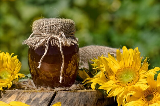 Glas mit Honig und Sonnenblumen auf Holztisch über Bokeh-Gartenhintergrund Glas mit frischem Honig im Feld der Wildblumen