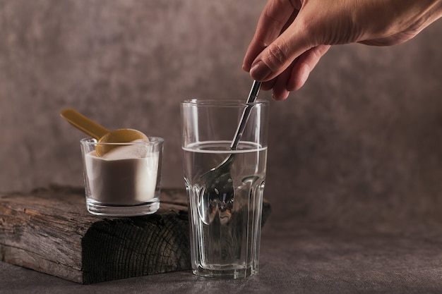 Glas mit in Wasser gelöstem Kollagen und Kollagenproteinpulver. Frauenhand hält einen Löffel. Gesundes Lebensstilkonzept.