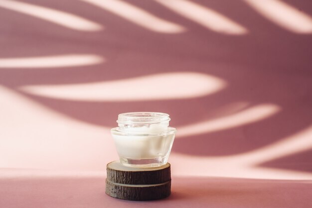 Glas mit weißer Feuchtigkeitscreme auf Holzsäge schneidet auf einem rosa Hintergrund. Lotion auf dem Hintergrund des tropischen Schattens eines Palmblatts. Das Konzept der Hautpflege