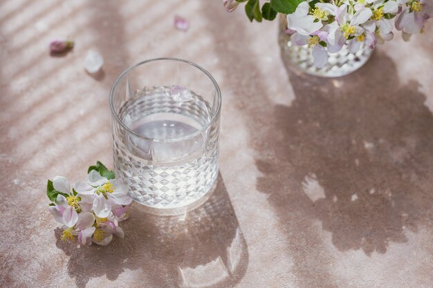 Glas reines Wasser auf dem Tisch mit blühendem Apfelbaumzweig in einem Glas. Morgensonne Stimmung