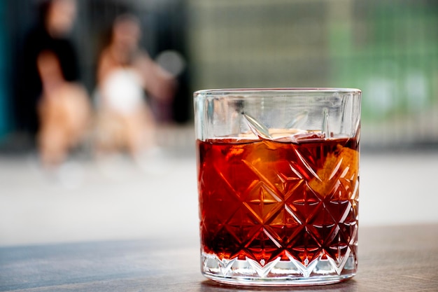 Glas roter Negroni-Cocktail mit Orangenscheiben und klarem Eisblock auf Holztischhintergrund verschwommene Stadt und Menschen