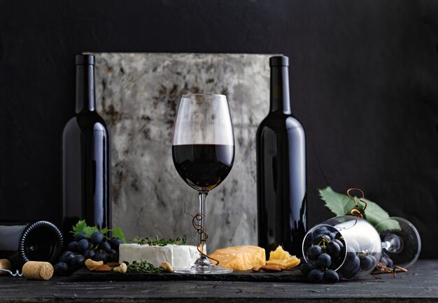 Glas rotwein mit snacks und käse auf dunklem hintergrund. glas und flaschen rotwein auf dunklem, schwermütigem schwarzem betonhintergrund.