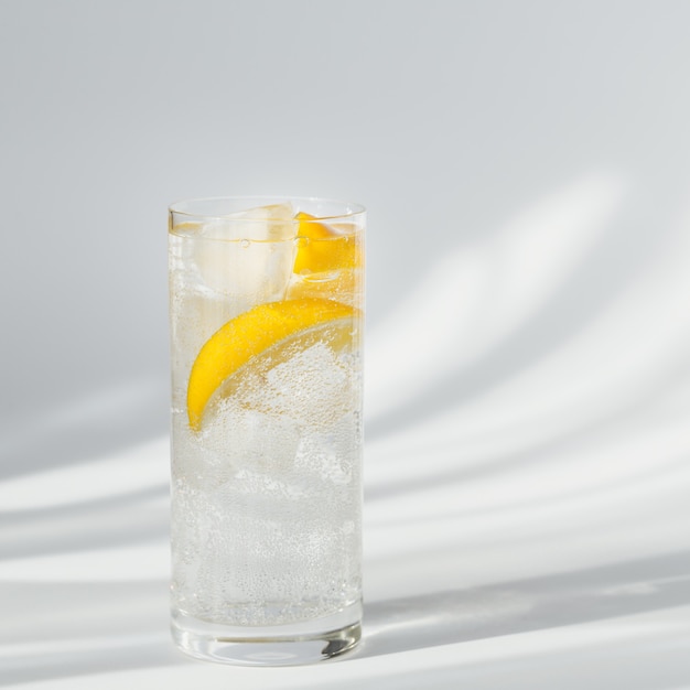 Glas sauberes Mineral-Sprudelwasser mit Eis und Zitrone auf weißem Hintergrund mit Sonnenschein. Licht mit harten Schatten und Blendung aus dem Glas. Frühstück, frisches Morgengetränk