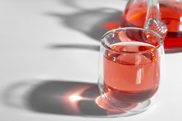 Glas Teekanne und Glas Tasse mit Tee auf weißem Hintergrund mit langen Schatten schließen