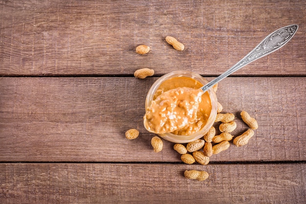 Glas und Löffel voll knuspriger Erdnussbutter mit Erdnüssen auf den Punkt gebracht auf Holz