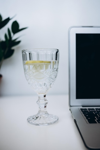 glas wasser mit zitronenlaptop und gelbem blumenstrauß auf weißem schreibtisch