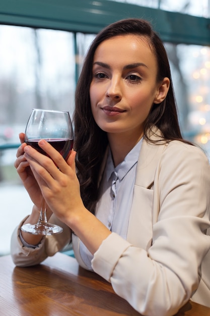 Glas Wein. Nette nachdenkliche gut aussehende Frau, die Glas Wein erhöht, während sie am Tisch sitzt und meditiert