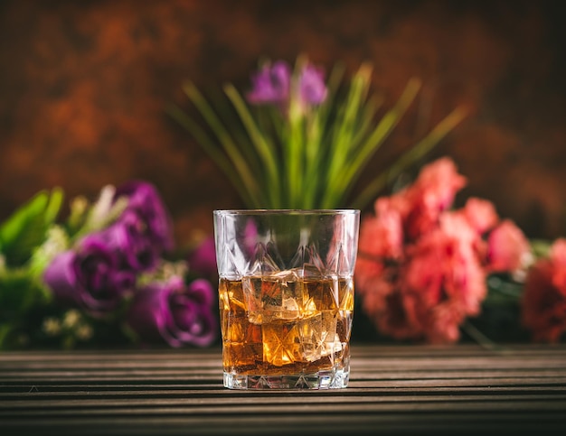 Glas Whiskey mit Eis auf einem Holztisch mit einigen Farbblumen