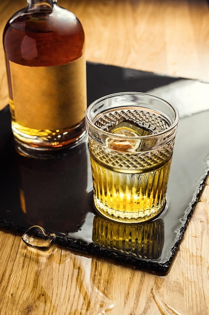 Glas Whisky Cognac oder Brandy mit Zitrone und Eiswürfeln, die auf der Theke mit einer Flasche im Hintergrund stehen