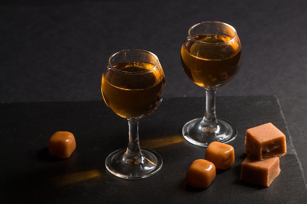 Glas Whisky mit Karamellbonbons auf einem schwarzen Steinschieferbrett auf schwarzem Hintergrund. Seitenansicht, zurückhaltend.