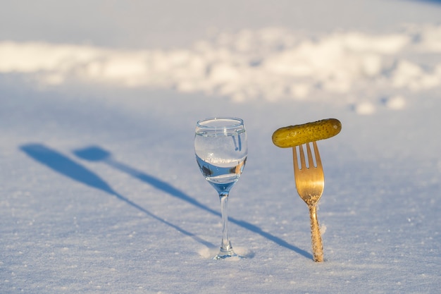 Glas Wodka und eine Gabel mit eingelegter Gurke auf weißem Schnee im Winter, Nahaufnahme, Ukraine