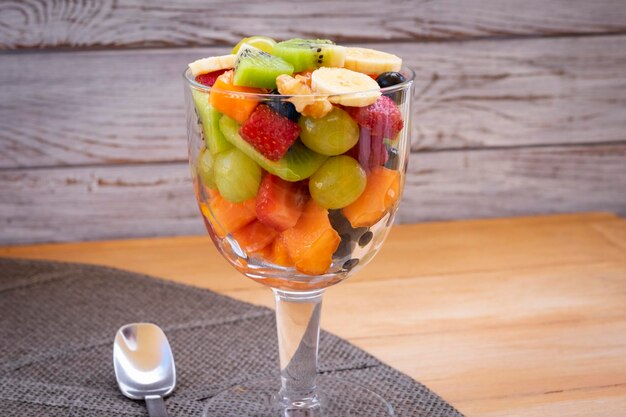 Glasbecher mit gesundem, frischem Obstsalat mit Trauben, Erdbeere, Papaya, Banane, Kiwi, Heidelbeeren