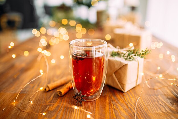 Glasbecher schwarzer Tee mit Sternanis auf dem hölzernen festlichen Tisch mit Girlandenlichtern