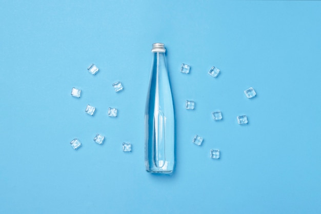 Glasflasche mit klarem Wasser auf einer blauen Oberfläche mit Eiswürfeln. Konzept von Gesundheit und Schönheit, Wasserhaushalt, Durst, Hitze, Sommer. Flachgelegt, Draufsicht.