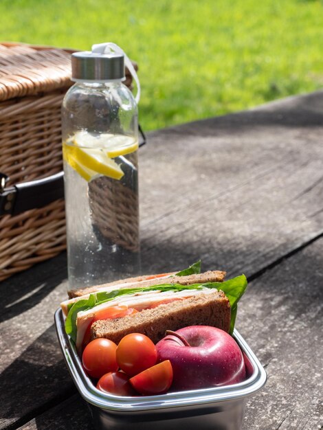 Glasflasche mit Wasser und Lunchbox aus Edelstahl mit Sandwich und Obst auf Holztischgras