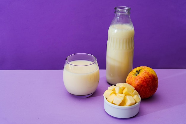 Glasflasche und großes Glas mit Milch und einem ganzen roten Apfel neben einer Schüssel mit Apfelwürfeln in einer violetten oder violetten Umgebung Kopieren Sie Platz