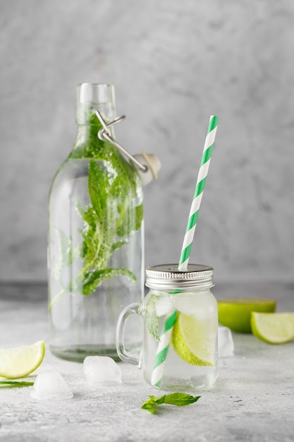 Glasflasche und Limonadenglas mit kalter Limonade mit frischen Minzblättern und Limette mit Eiswürfeln auf grauem Beton