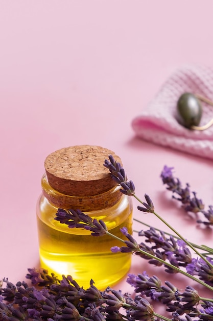 Glasölflasche mit Lavendelblüten auf farbigem Hintergrund mit Gesichtsmassagegerät im Hintergrund