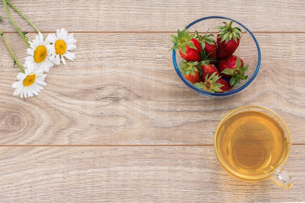 Glastasse grüner Tee, Kamillenblüten, Glasschüssel mit frischen Erdbeeren auf dem Holzhintergrund. Draufsicht mit Kopienraum.