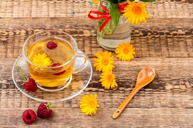Glastasse grüner Tee mit Ringelblumenblüten und frischen Himbeeren auf Holzhintergrund. Gesundes Getränk.