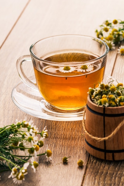Glastasse grüner Tee mit weißen Kamillenblüten und kleinem Holzfass mit trockenen Kamilleblüten im Vordergrund. Geringe Schärfentiefe. Konzentrieren Sie sich auf eine Tasse Tee