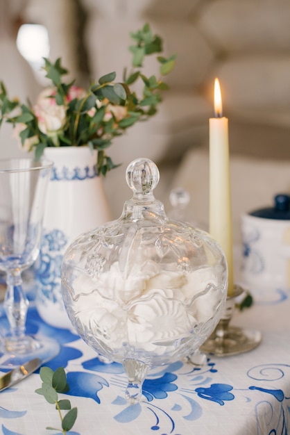 Glasvase mit Dessert Marshmallow ist auf dem Tisch, mit einer Tischdecke mit einem Muster Gzhel bedeckt. Neben der Kerze brennt. Dekor für ein festliches Abendessen oder Mittagessen